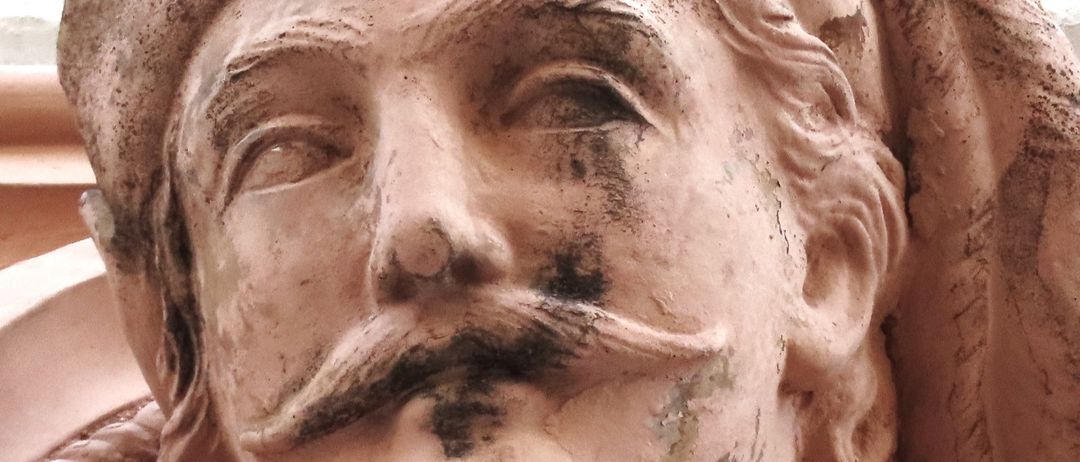 Eine Nahaufnahme einer verwitterten Statue, die einen bärtigen Mann mit Schnurrbart zeigt, der einen Hut trägt und auf seiner Oberfläche feine Details aufweist.