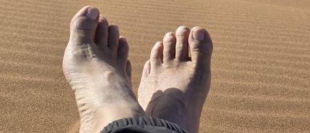 Die Füße einer Person liegen auf dem Sand mit einem Kamel im Hintergrund. Das Kamel läuft in der Wüste.