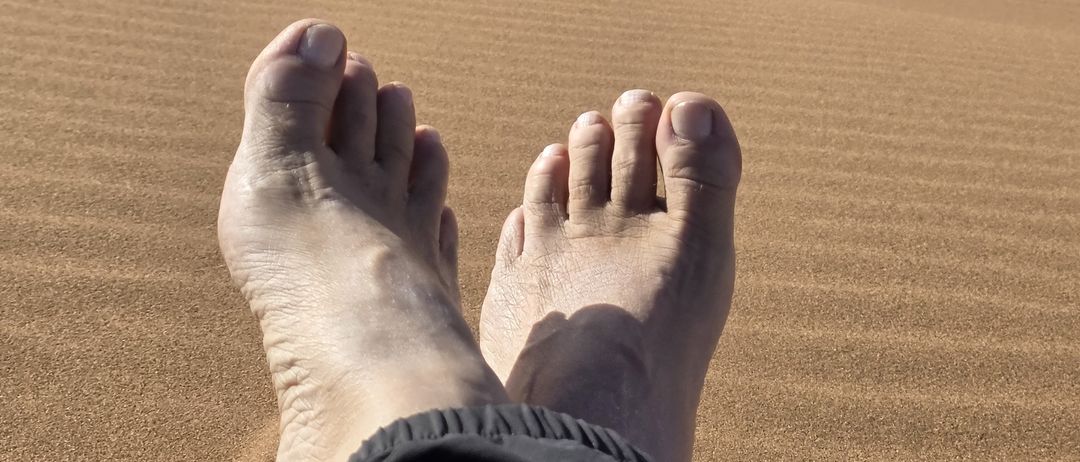 Die Füße einer Person liegen auf dem Sand mit einem Kamel im Hintergrund. Das Kamel läuft in der Wüste.
