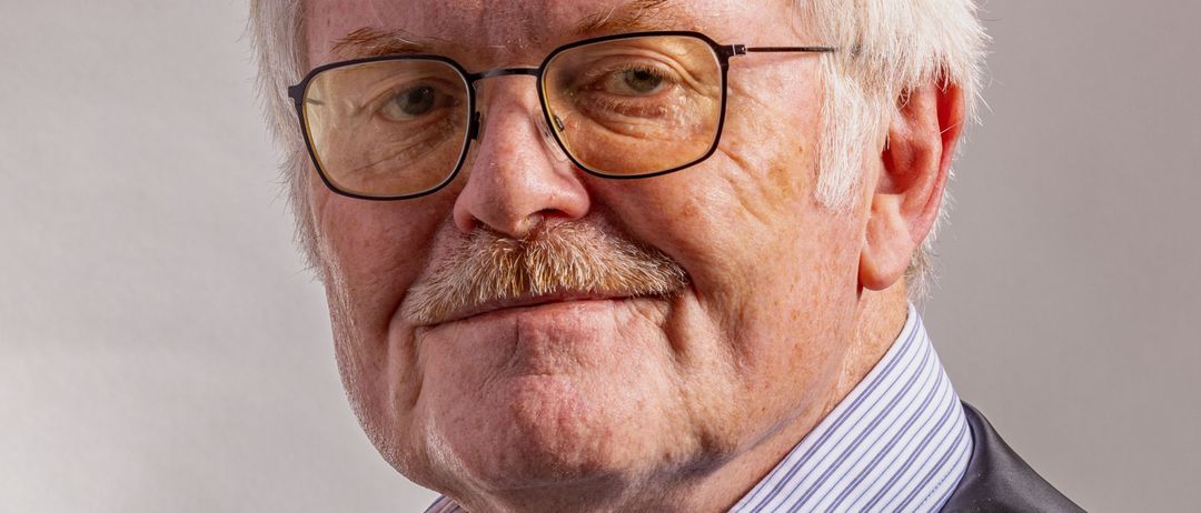 Ein älterer Mann mit weißem Haar und einem Schnurrbart, der eine Brille und eine graue Weste über einem blau gestreiften Hemd trägt.