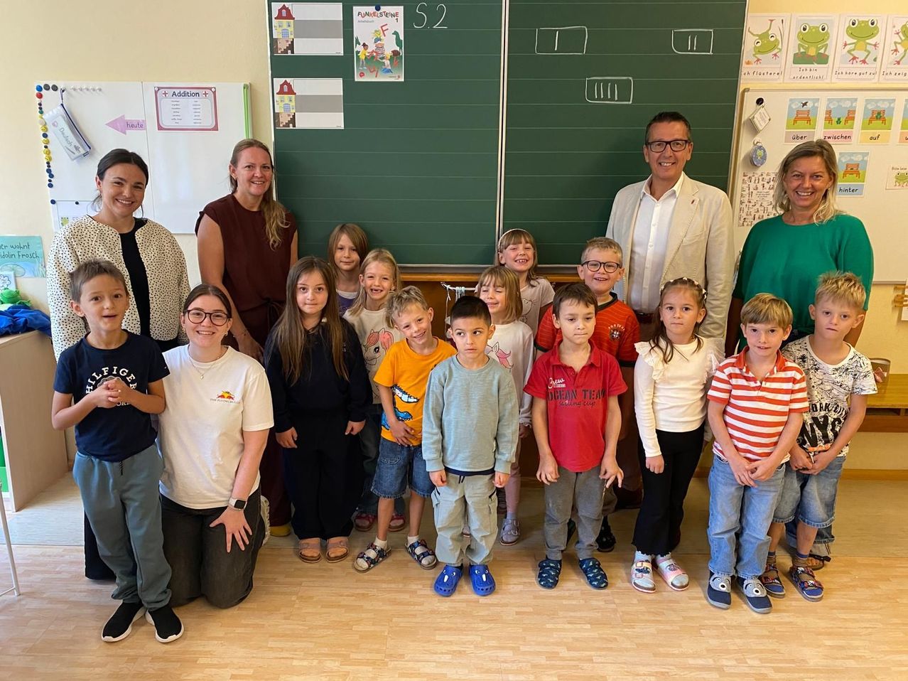 A group of children and adults are posing for a photo in a classroom. The children are standing in front of a blackboard with drawings and text.