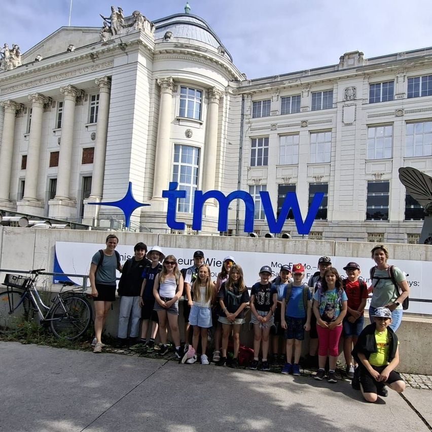 Eine Gruppe junger Leute mit Rucksäcken und Mützen steht vor einem Gebäude mit vielen Fenstern und einem blauen Sternenlogo.