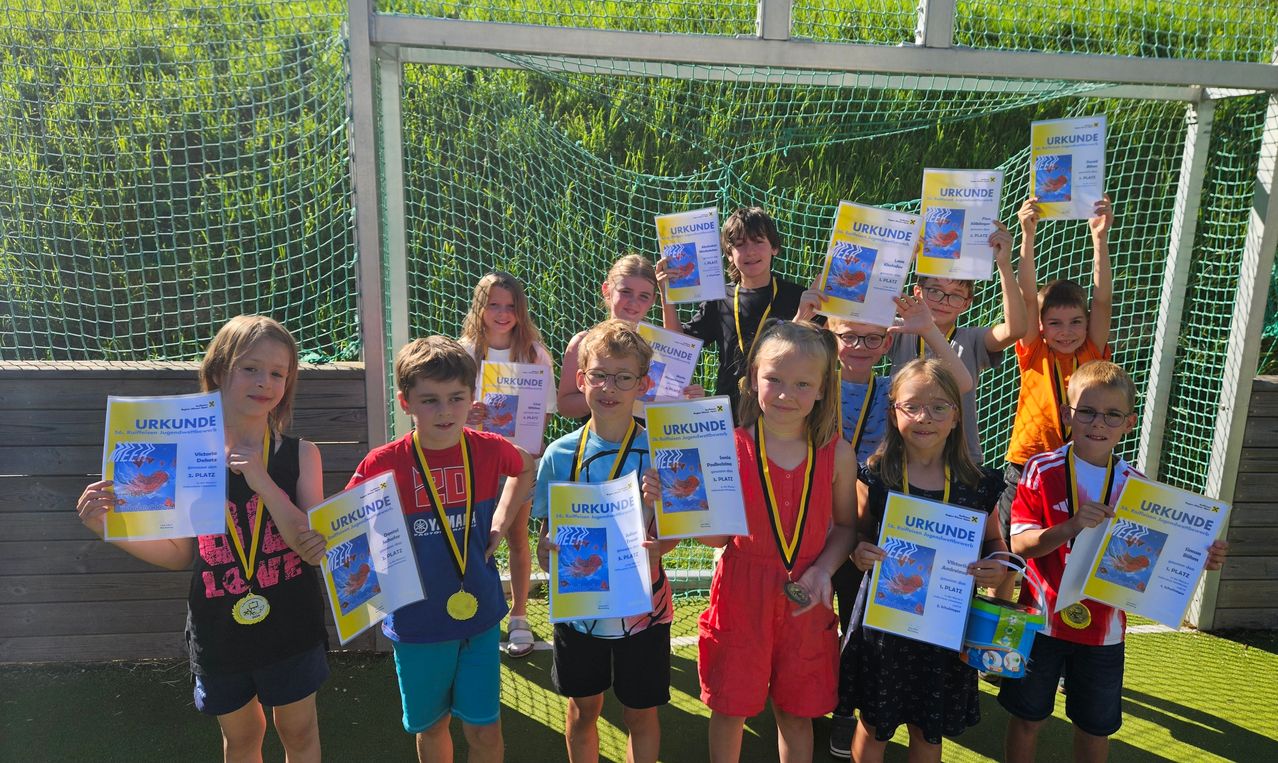 Eine Gruppe von Kindern mit Medaillen um den Hals halten Urkunden mit dem Wort 'Urkunde' hoch. Sie stehen auf einem Feld mit einem Netz im Hintergrund.