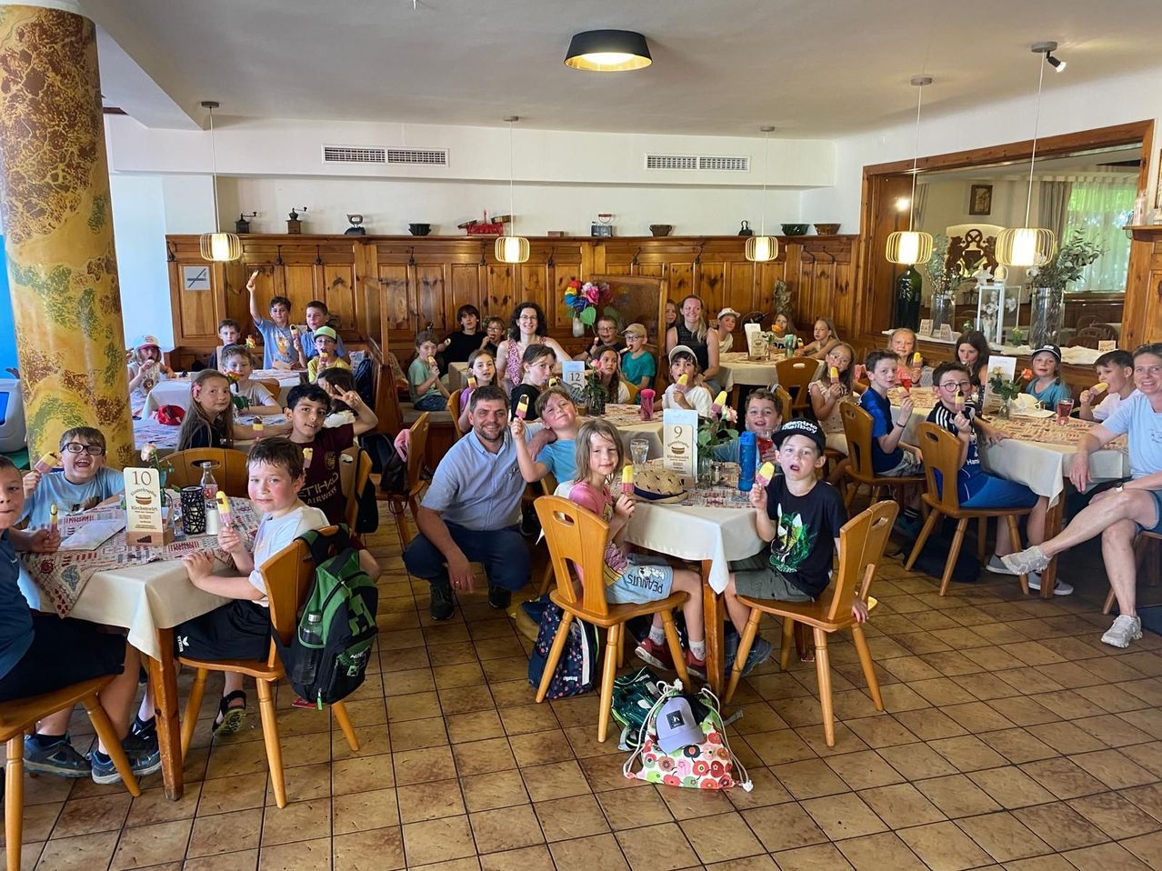 Eine Gruppe von Kindern und Erwachsenen in einem Esszimmer feiert mit Eis. Ein Mann hockt lächelnd. Holzstühle und Tische mit weißen Tischdecken sind zu sehen.
