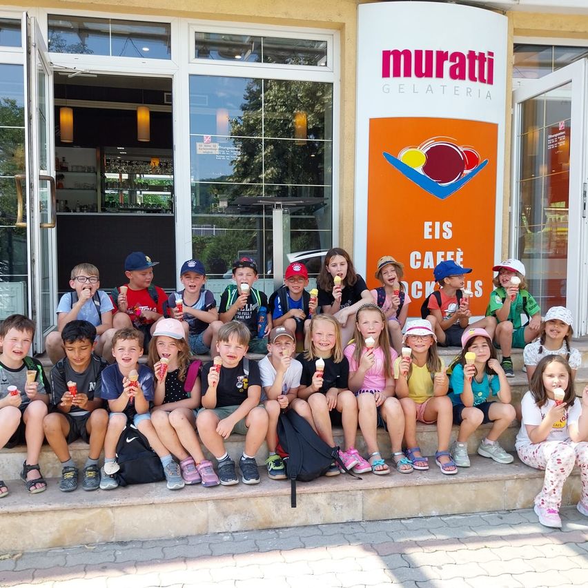 A group of children sit on the steps outside a gelateria, holding ice creams. They are in front of a building with a sign that says Muratti Gelateria.