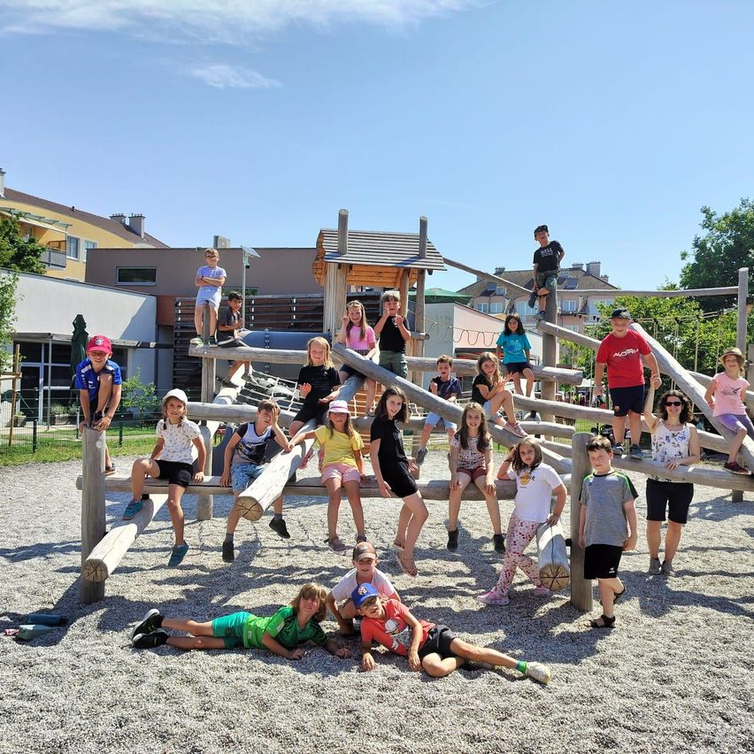A group of children and adults are posing for a photo in a playground with wooden structures. Some are standing, sitting, and lying on the ground. Trees and buildings are in the background.