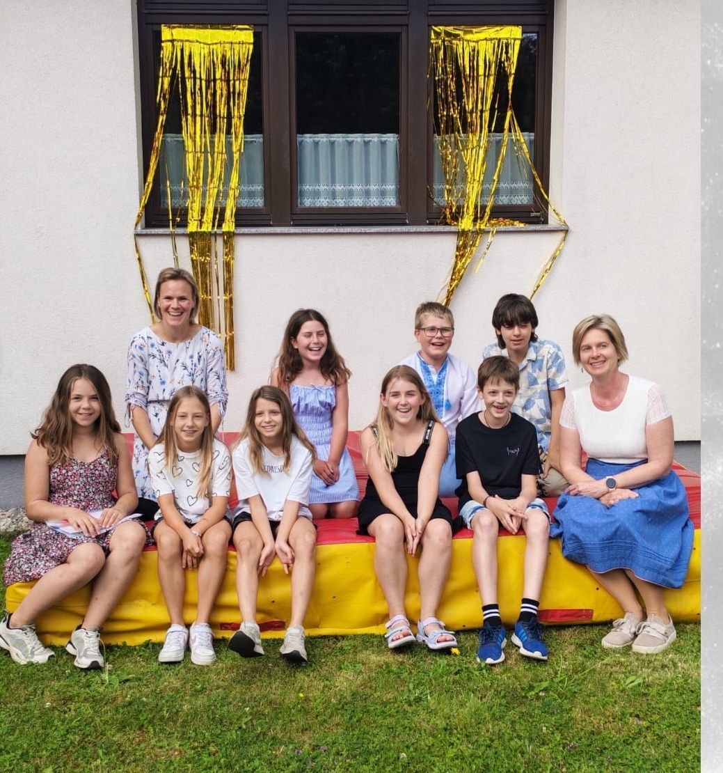 A group of children and two women pose for a photo on a yellow mat outside a building with a window and yellow streamers.