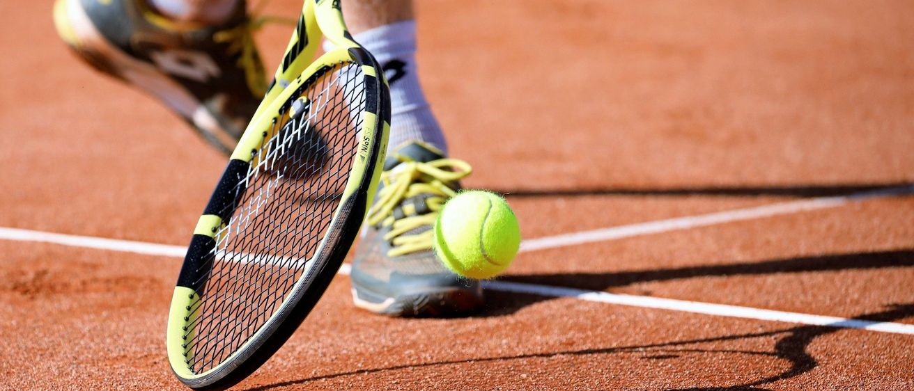 Der Fuß und der Schläger eines Tennisspielers treffen einen Tennisball auf einem Tennisplatz mit Schatten auf dem Boden.