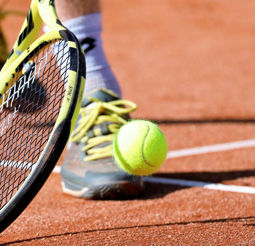 Der Fuß und der Schläger eines Tennisspielers treffen einen Tennisball auf einem Tennisplatz mit Schatten auf dem Boden.