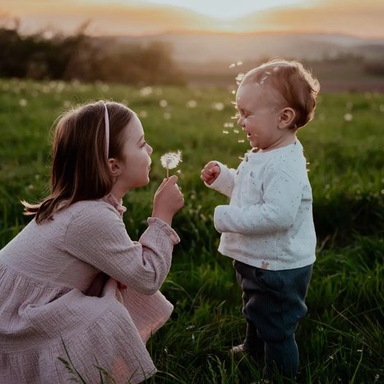 Ein junges Mädchen hält ein Löwenzahn und pustet darauf, während ein Baby in einem Grasfeld unter einem Sonnenuntergangshimmel lacht.