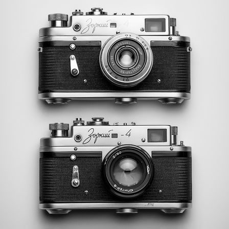 Zwei Vintage-Kameras sind ausgestellt, eine mit einem Objektiv, das 'Zenit-4' und '2.8/50' zeigt, die andere mit einem Objektiv, das '25/50' und '2/50' zeigt. Beide Kameras haben strukturierte schwarze und silberne Korpusse.