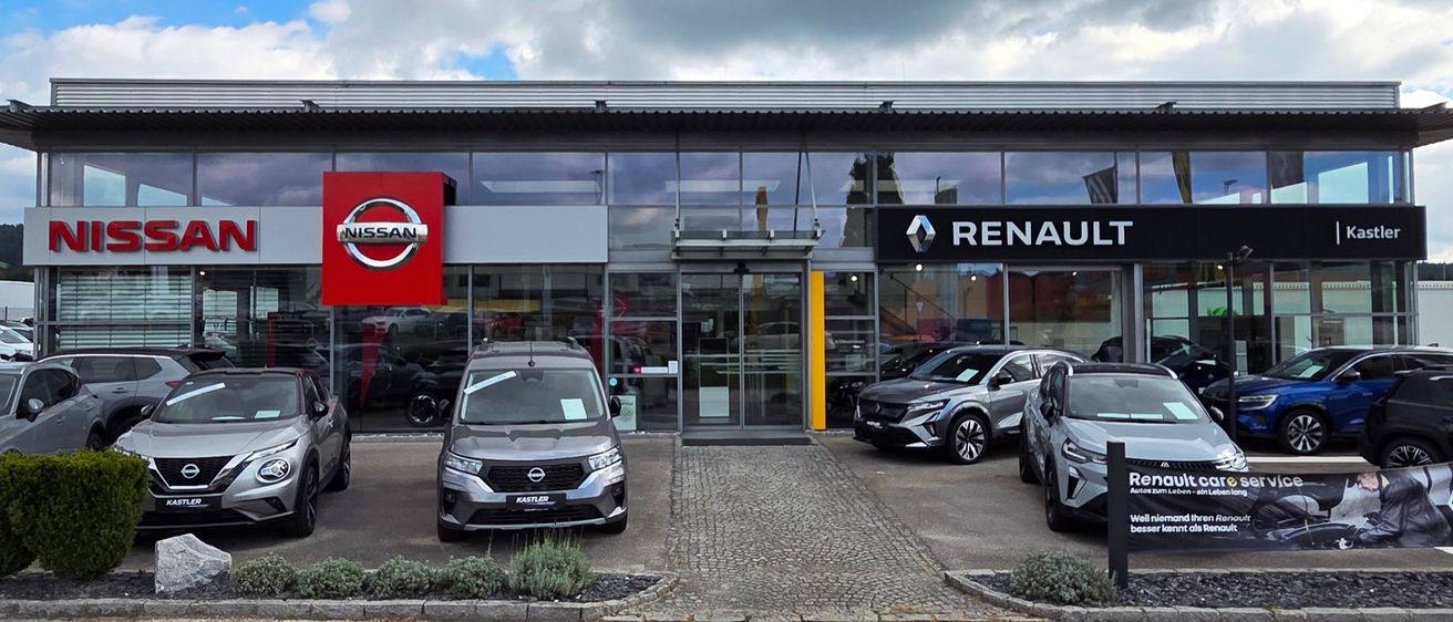 Ein Nissan und ein Renault Auto sind vor einem Händler mit Glaswänden geparkt, der sich unter einem bewölkten Himmel befindet.