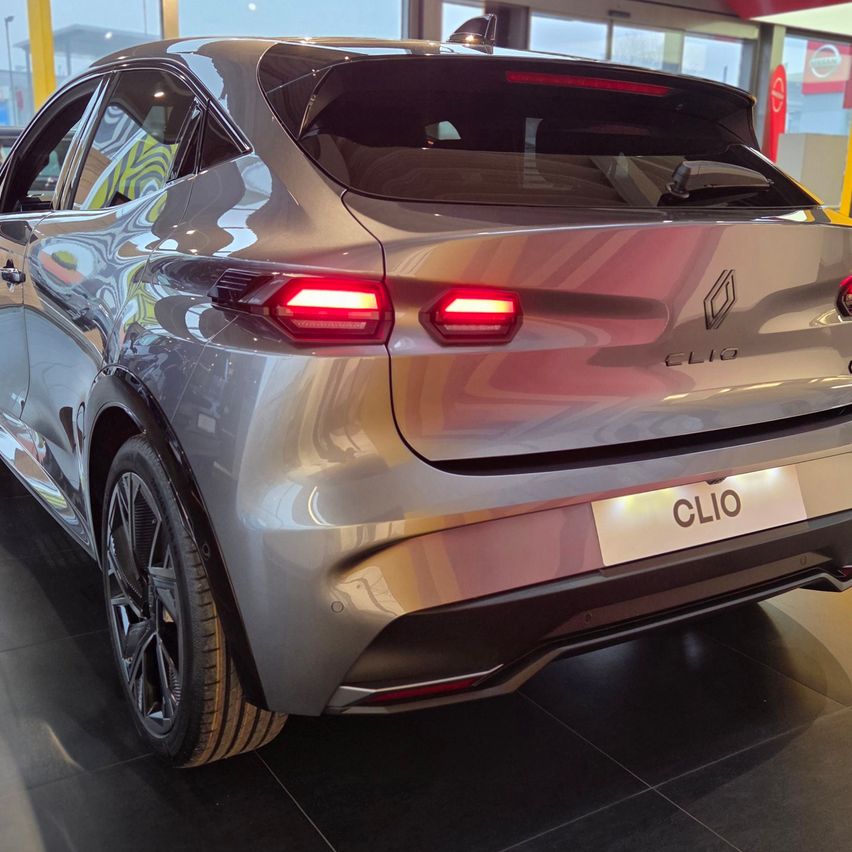 Die Rückseite eines silbernen Renault Clio ist in einem Ausstellungsraum mit schwarzen Fliesenboden ausgestellt. Das Auto hat ein schlankes Design und rote Rückleuchten.