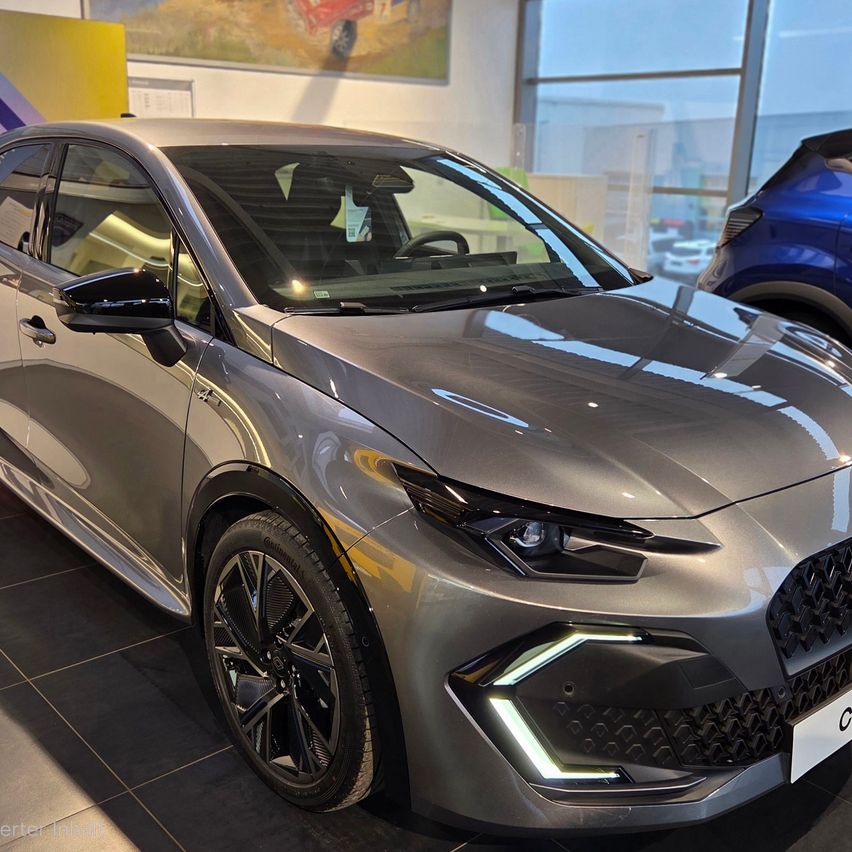 Ein metallisch graues Auto mit schlanken Scheinwerfern und einem modernen Kühlergrill steht in einem Autohaus. Das Auto hat ein schlankes Design und ist von anderen Fahrzeugen umgeben.