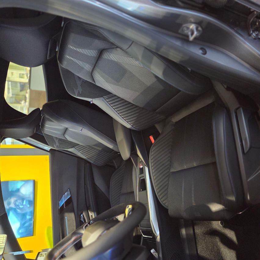 Der Innenraum eines Autos zeigt graue Ledersitze und ein Lenkrad. Die Sitze sind gut gepolstert mit sichtbaren Nähten. Das Armaturenbrett hat Bedienknöpfe und eine Lüftung.