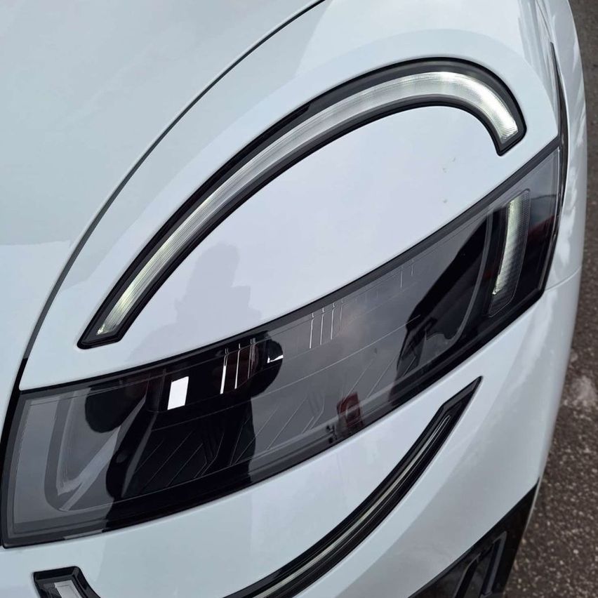 Eine Nahaufnahme der Front eines weißen Autos zeigt seine Scheinwerfer. Die Scheinwerfer haben ein modernes Design mit LED-Beleuchtung. Der Stoßfänger des Autos hat ein schlankes Aussehen.