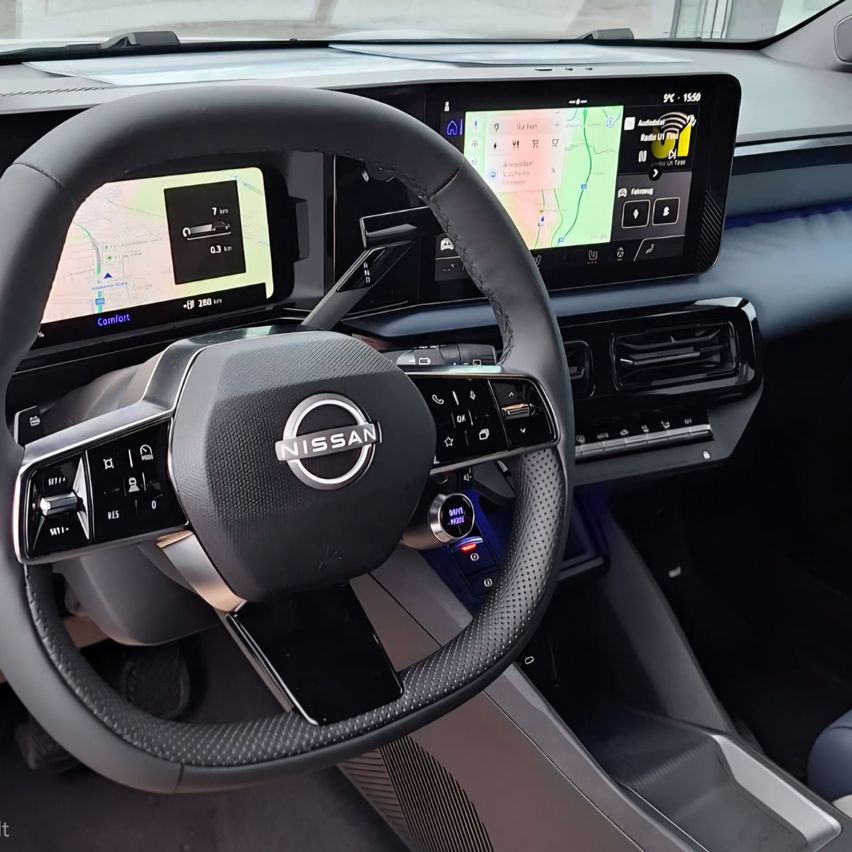 Das Armaturenbrett eines Nissan-Fahrzeugs zeigt ein digitales Display mit Karte, Navigation und Fahrzeugeinstellungen. Das schwarze Lenkrad hat ein silbernes Nissan-Logo.