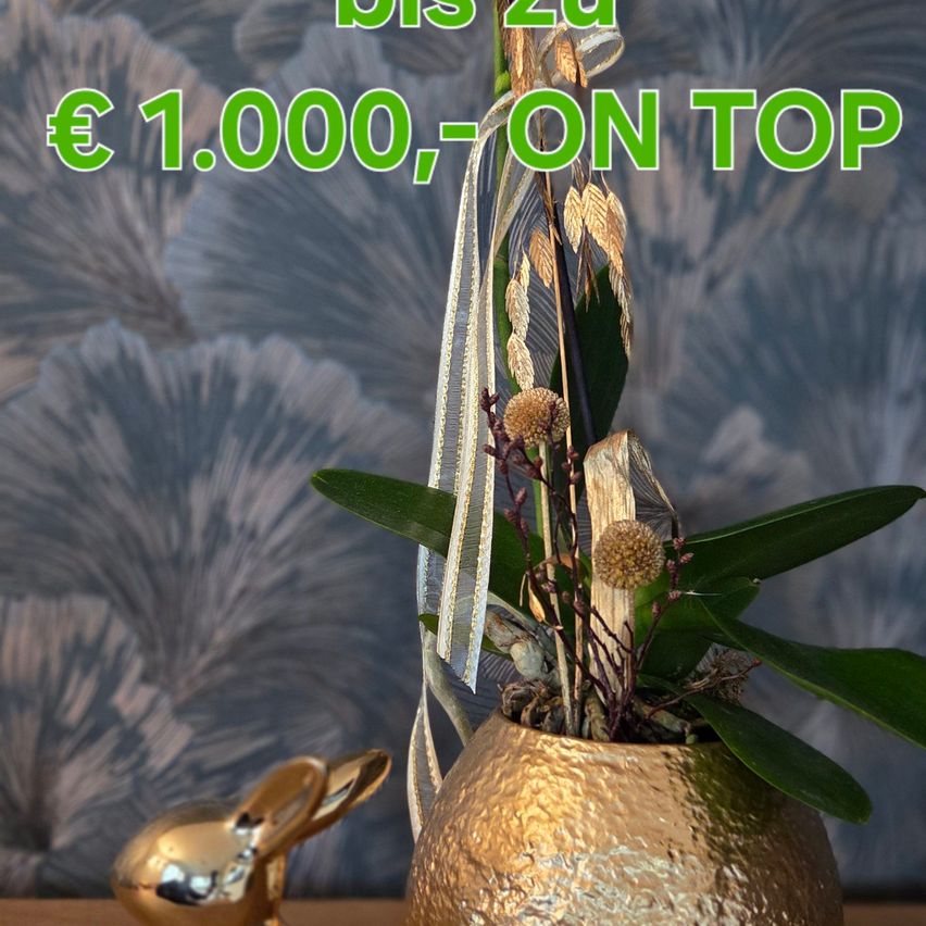 Eine goldene Topfpflanze mit grünen Blättern und einer goldenen Schleife steht auf einem Tisch mit einem goldenen Hasen. Der Text darüber lautet 1.000€, Obendrauf.