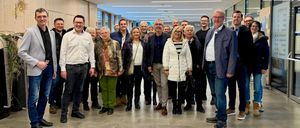 Eine Gruppe von Menschen, wahrscheinlich Kollegen, posiert für ein Foto in einem modernen Flur. Sie stehen in zwei Reihen, wobei einige Personen eine Brille tragen. Der Flur hat weiße Wände, geflieste Böden und Deckenleuchten. Eine Uhr ist an der Wand montiert.