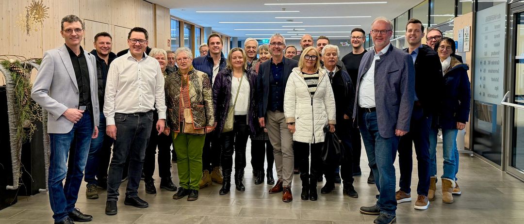 Eine Gruppe von Menschen, wahrscheinlich Kollegen, posiert für ein Foto in einem modernen Flur. Sie stehen in zwei Reihen, wobei einige Personen eine Brille tragen. Der Flur hat weiße Wände, geflieste Böden und Deckenleuchten. Eine Uhr ist an der Wand montiert.