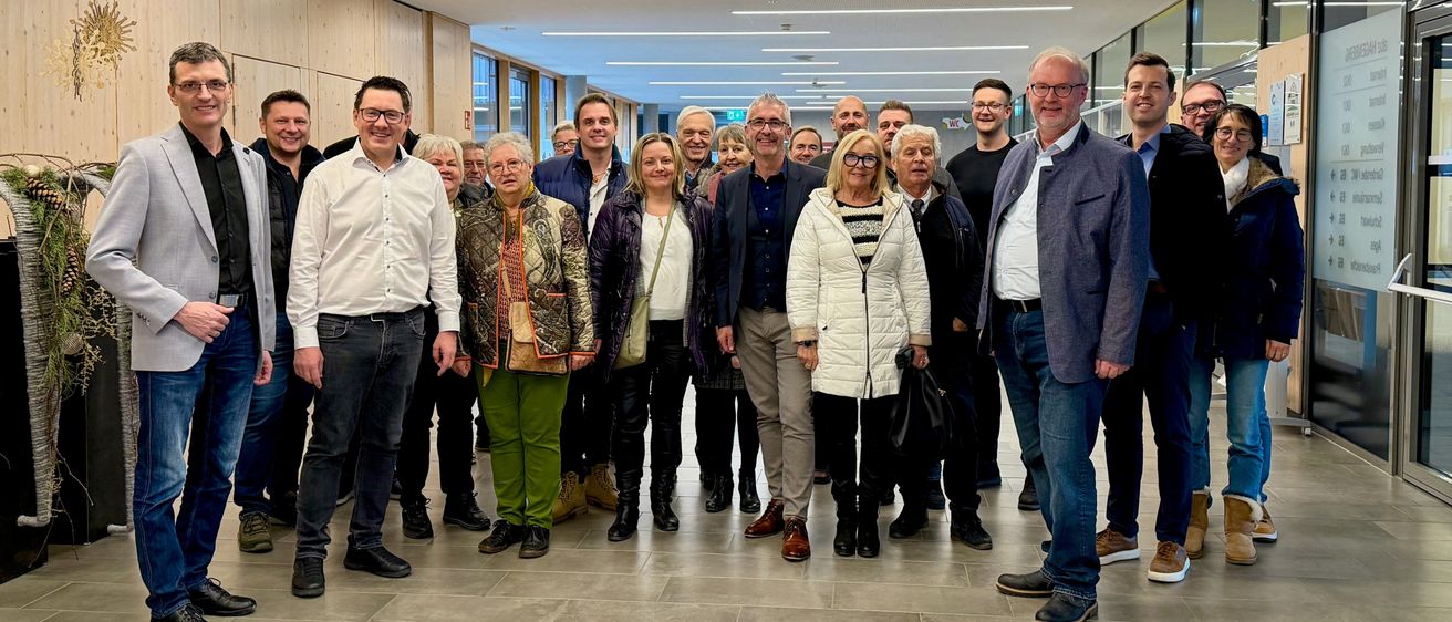 Eine Gruppe von Menschen, wahrscheinlich Kollegen, posiert für ein Foto in einem modernen Flur. Sie stehen in zwei Reihen, wobei einige Personen eine Brille tragen. Der Flur hat weiße Wände, geflieste Böden und Deckenleuchten. Eine Uhr ist an der Wand montiert.
