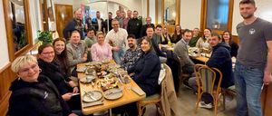 Eine Gruppe von Menschen versammelt sich um einen Tisch in einem Restaurant und lächelt für ein Foto. Sie tragen Winterkleidung und sitzen auf Stühlen. Der Tisch hat Teller, Tassen und Schüsseln. Ein Mann steht hinter dem Tisch. Der Raum hat weiße Wände, Holzbalken und Spiegel.