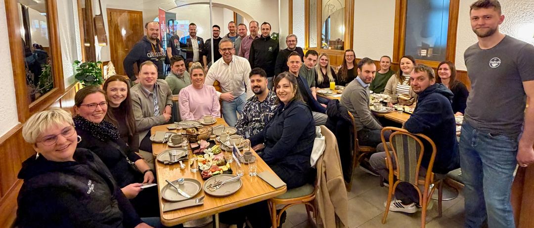 Eine Gruppe von Menschen versammelt sich um einen Tisch in einem Restaurant und lächelt für ein Foto. Sie tragen Winterkleidung und sitzen auf Stühlen. Der Tisch hat Teller, Tassen und Schüsseln. Ein Mann steht hinter dem Tisch. Der Raum hat weiße Wände, Holzbalken und Spiegel.