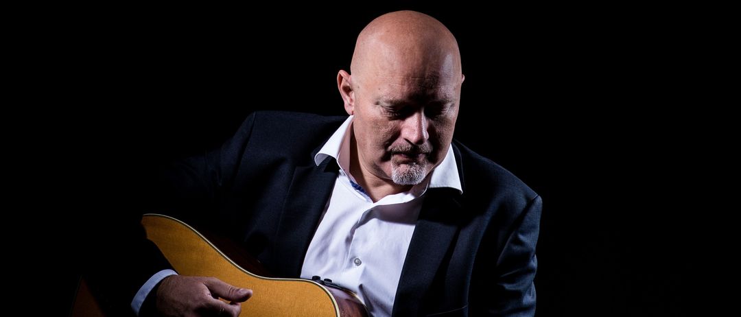 Ein kahlköpfiger Mann spielt Gitarre in einem dunklen Raum. Er trägt ein weißes Hemd und einen schwarzen Blazer. Er scheint konzentriert zu spielen.