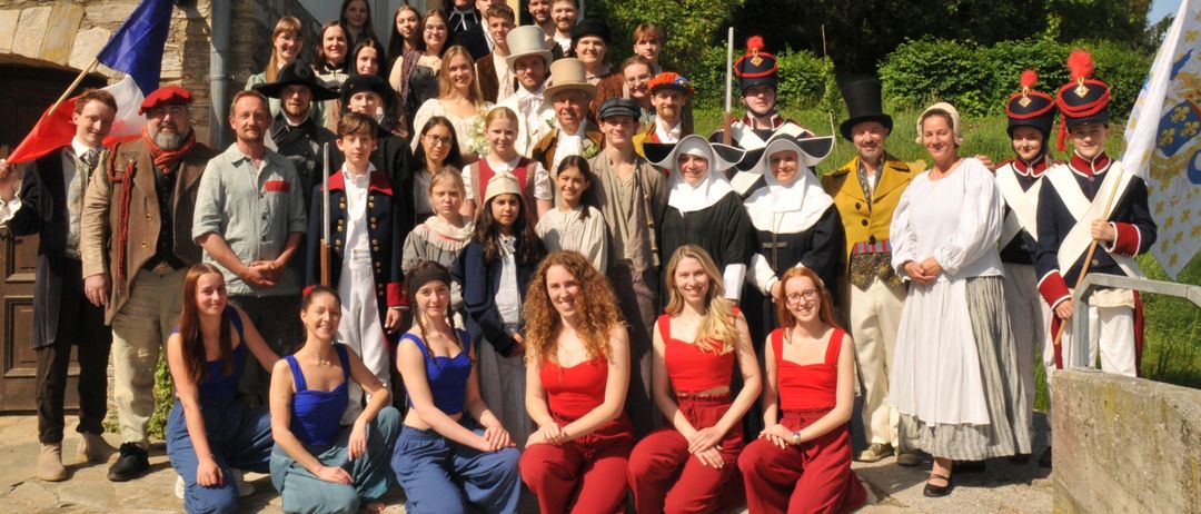 Eine Gruppe von Menschen in historischen Kostümen posiert für ein Foto, einige in roten Outfits, andere in verschiedenen historischen Gewändern, mit Grünpflanzen und einem Gebäude im Hintergrund.