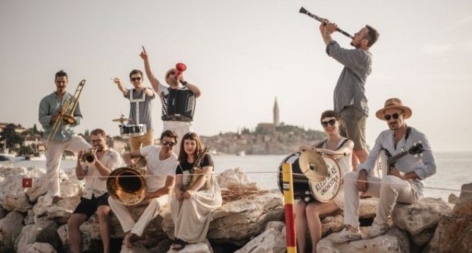Eine Gruppe Musiker versammelt sich auf Felsen am Wasser, einige spielen Instrumente, andere stehen oder sitzen, mit einer Stadtsilhouette im Hintergrund.