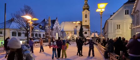 Bei Nacht ist eine Eisbahn voller Menschen, einige skatend und andere zuschauend. Hohe Straßenlaternen und Gebäude im Hintergrund.