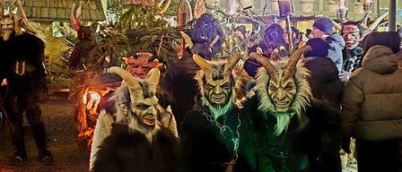 Menschen in Hörnermasken stehen nachts vor einer dekorierten Straße. Dahinter sind Gebäude mit Weihnachtsbeleuchtung und eine Menschenmenge zu sehen.