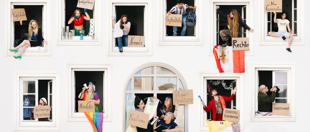 Eine Gruppe von Menschen hält Schilder in verschiedenen Fenstern eines Gebäudes. Einige Schilder enthalten 'Moralistin', 'Regie-Assistenz' und 'Kostüme'. Ein Mann und eine Frau stehen am Eingang.