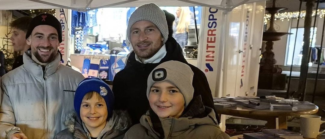 Eine Gruppe von Menschen steht unter einem Zelt. Sie lächeln und posieren für ein Foto. Auf einem Holztisch liegen Bücher und ein Stift. Dahinter stehen weitere Menschen, einige stehen in der Nähe einer weißen Wand.