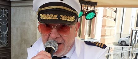 Ein Mann in einer Kapitänsuniform singt unter einem Zelt in ein Mikrofon. Dahinter befindet sich ein Gebäude mit grünen Lichtern.