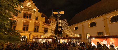 Ein geschäftiger Weihnachtsmarkt vor einem großen Gebäude mit Lichterketten. Viele Menschen sind versammelt, einige in Winterkleidung. Das Gebäude hat viele Fenster und Dekorationen.
