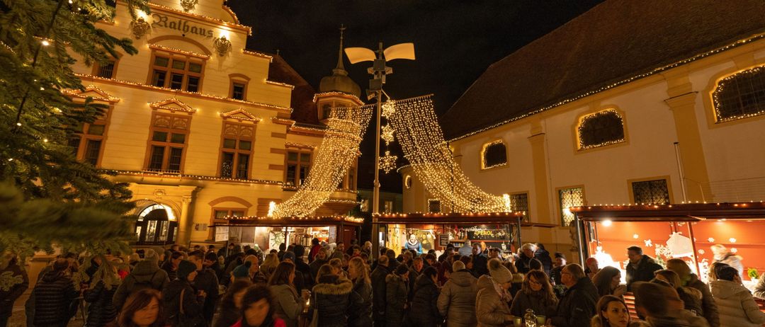 Ein überfüllter Außenbereich bei Nacht mit festlichen Lichtern, Menschen in Winterkleidung und einem Gebäude mit der Aufschrift Rathaus.