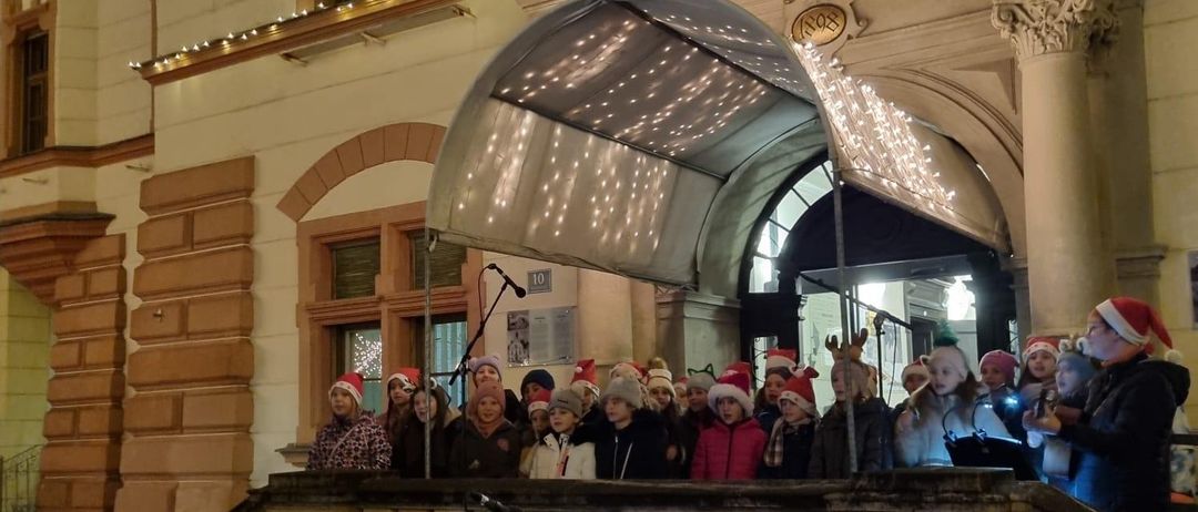 Ein Chor von Kindern mit Weihnachtsmützen tritt auf den Stufen eines dekorierten Gebäudes unter einem beleuchteten Vordach bei Nacht auf.