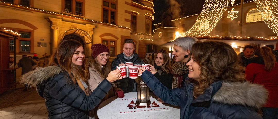 Eine Gruppe von Menschen feiert um einen Tisch mit beleuchteten Bechern unter einem Nachthimmel mit Stadtlichtern.