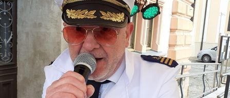 Ein Mann in einer weißen Uniform mit goldenen Streifen und einer Kapitänsmütze hält ein Mikrofon und singt, möglicherweise bei einer Veranstaltung.