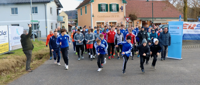 Eine Gruppe von Menschen in blauer und grauer Sportbekleidung läuft auf einer Straße. Ein Mann macht ein Foto. Ein Haus und ein Banner sind auf der linken Seite.