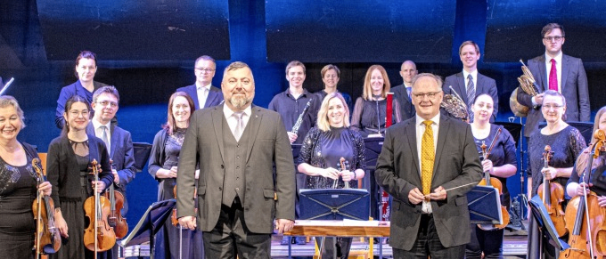 Eine Gruppe von Musikern, darunter Geiger, steht auf der Bühne mit einem Dirigenten. Die Geiger halten ihre Instrumente und der Dirigent hält einen Taktstock. Die Bühne ist mit Blumen und Musikinstrumenten geschmückt.