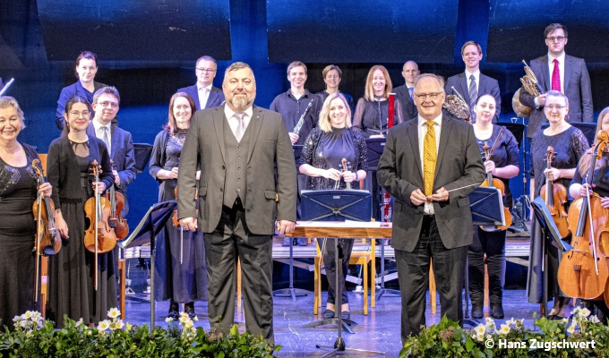 Eine Gruppe von Musikern in Anzügen und Krawatten steht mit Musikinstrumenten auf der Bühne. Zwei Männer stehen vorne, einer hält einen Dirigentenstab.