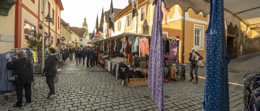 A busy street market with numerous stalls displaying various clothing items. Flags hang above the stalls, and people are walking on the cobblestone street.