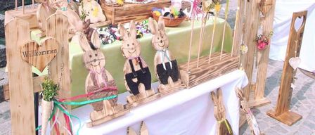 Eine hölzerne Ausstellung von geschnitzten Häschen auf einem Tisch, geschmückt mit bunten Bändern und Herzzeichen. Der Tisch hat eine weiße Tischdecke.