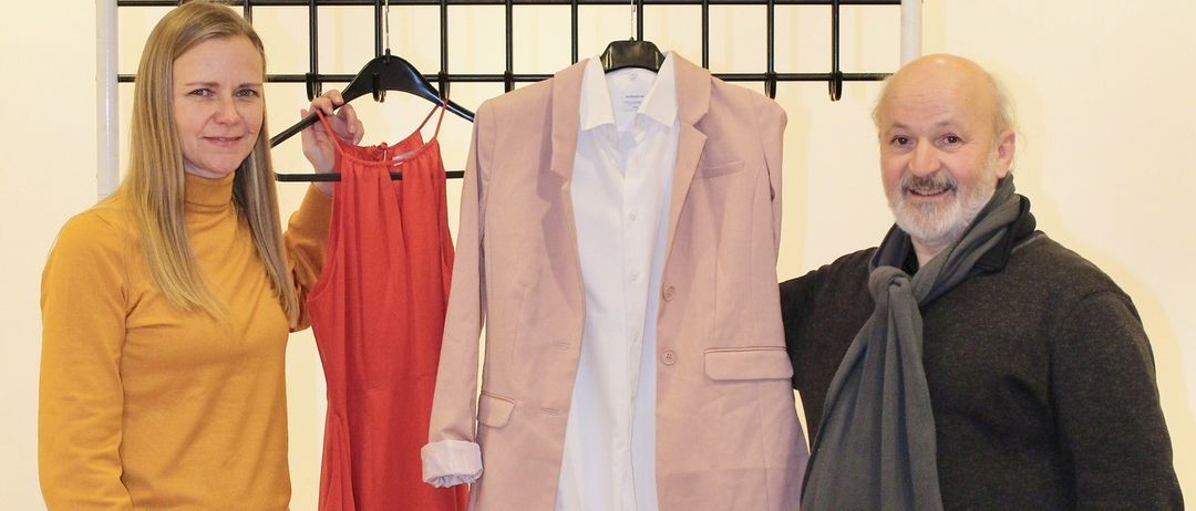 Zwei Personen stehen neben einem Tisch mit Kleidung. Eine Frau hält ein rotes Kleid. Ein Mann lächelt und trägt einen Schal.