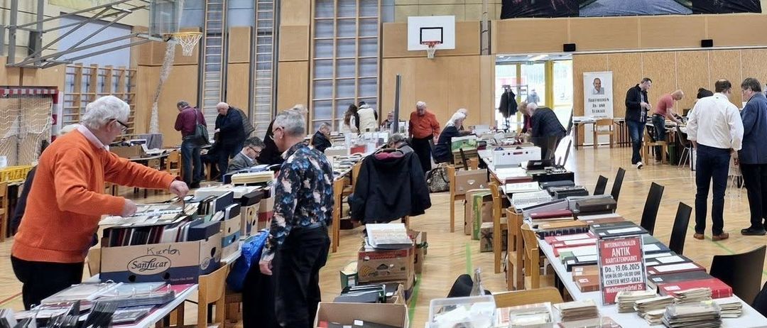 Im Inneren durchsuchen Menschen Gegenstände auf Tischen in einer Halle. Mehrere Tische sind aufgestellt, mit Büchern, Kisten und anderen Artikeln zur Ansicht. Die Leute schauen sich die Artikel an und heben sie auf. Die Halle hat einen Basketballkorb, Poster und ein Banner an der Wand.