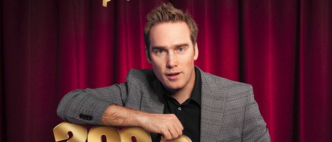 Ein Mann in einem Anzug steht vor einem roten Vorhang und hält ein goldenes Schild mit der Aufschrift '2002 bis Heute'. Er posiert für ein Foto.