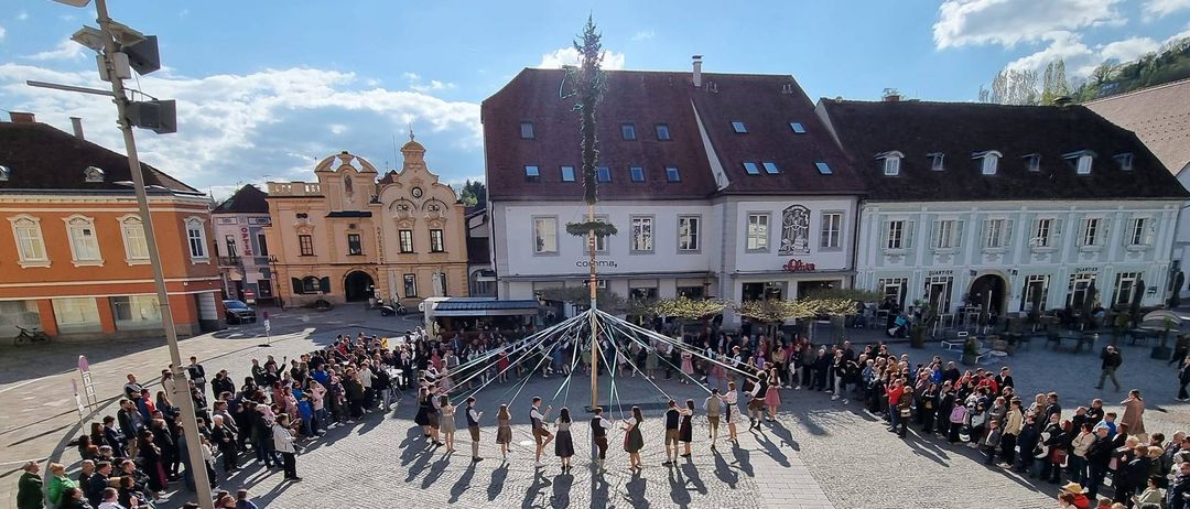 A crowd gathers around a maypole in a town square, with people walking and standing, buildings around, and a clear sky with clouds.