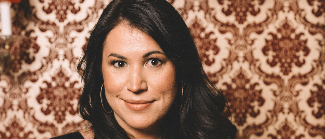Eine Frau mit schwarzen Haaren lächelt in die Kamera. Sie trägt ein schwarzes Oberteil. Hinter ihr ist eine gemusterte Tapete mit einem braun-beigen Design zu sehen. An der Wand links ist eine orangefarbene Lampe montiert.